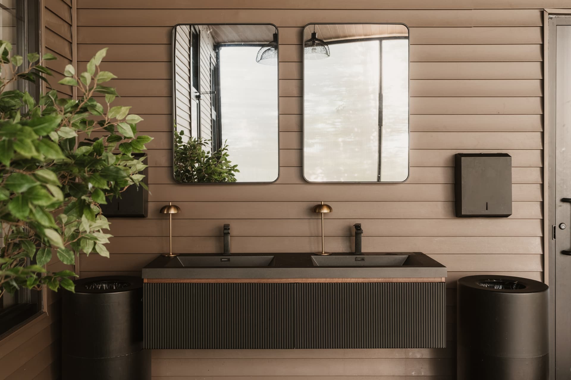 Double black basin vanity with paired mirrors, brass lamps and wood-panelled walls