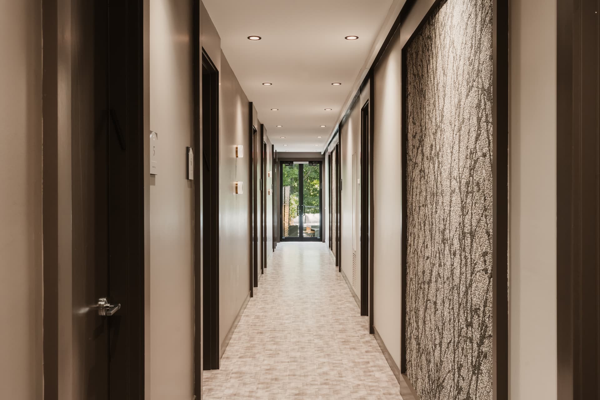Long guest room hallway with recessed lighting, textured wall panels and a view of the outdoor terrace